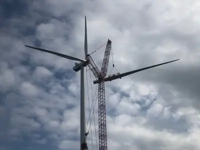 A crane is lifting equipment near the blades of a wind turbine against a cloudy sky.