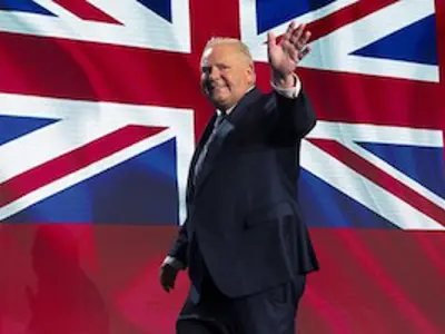 A person in a suit waving, with a large British flag in the background.
